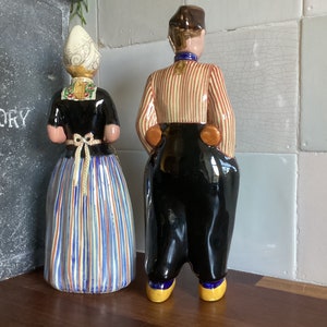 May include: A pair of ceramic figurines depicting a man and woman in traditional Dutch clothing. The man is wearing a striped shirt and black pants, while the woman is wearing a blue and white striped skirt and a black bodice. Both figurines have painted details and are standing on a wooden surface.