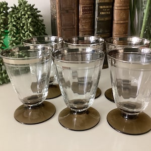 May include: Set of six clear glass wine glasses with a silver rim and a dark brown base. The glasses are arranged in a semi-circle on a white surface.