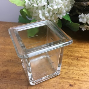 Op de afbeelding: Een vierkante glazen container met een dikke rand en gegradueerde markeringen. De container is leeg en staat op een houten oppervlak.