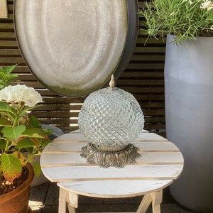 Può includere: Un paralume in vetro trasparente con un motivo a diamante poggia su una base argentata. Il paralume è su un tavolo di legno bianco con una finitura invecchiata.