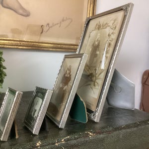 May include: Four silver picture frames of varying sizes, arranged in a row on a wooden surface. The frames are all ornate and have a vintage look. The largest frame is on the right and contains a black and white photo of two people. The other frames contain black and white photos of a single person.