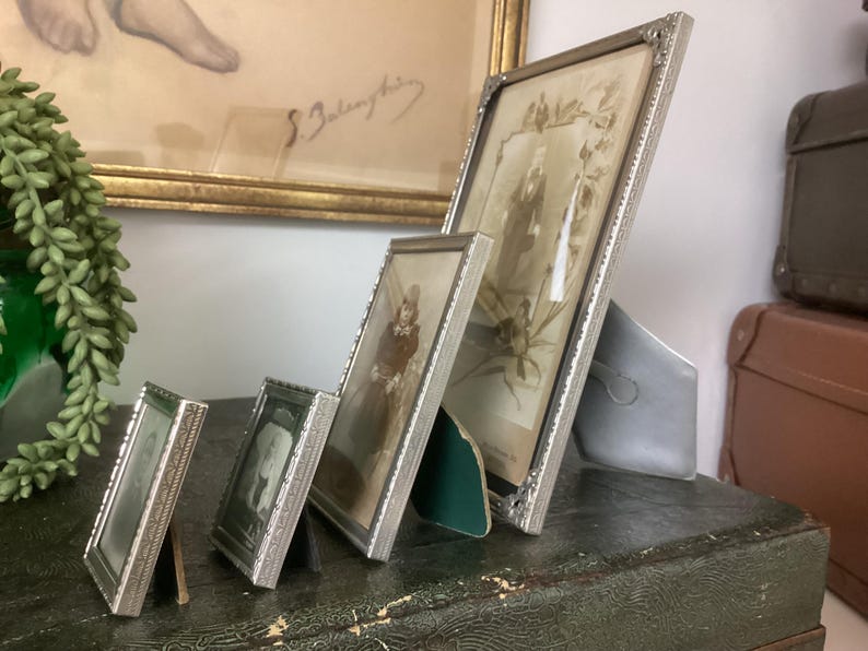 May include: Four silver picture frames of varying sizes, arranged in a row on a wooden surface. The frames are all ornate and have a vintage look. The largest frame is on the right and contains a black and white photo of two people. The other frames contain black and white photos of a single person.