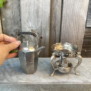 Peut inclure: Une petite théière argentée avec une surface texturée et une poignée noire, à côté d'un chauffe-plat argenté. La théière est tenue par une main. Le support a des pieds ornés et un dessus décoratif.