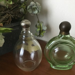 May include: Two vintage glass perfume bottles with metal stoppers. The bottle on the left is clear glass with a silver stopper. The bottle on the right is green glass with a brown stopper.