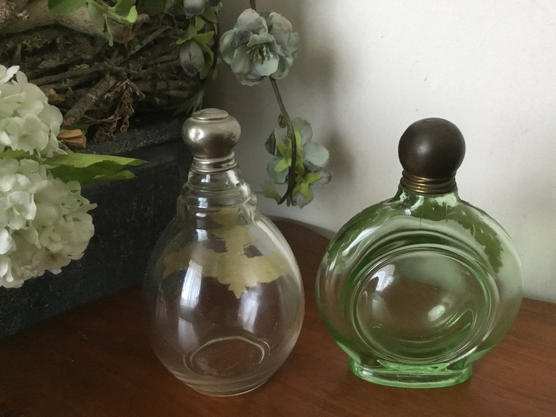 May include: Two vintage glass perfume bottles with metal stoppers. The bottle on the left is clear glass with a silver stopper. The bottle on the right is green glass with a brown stopper.