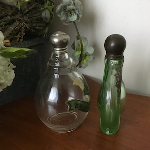 May include: Two vintage glass perfume bottles with silver tops. The bottle on the left is clear glass and has a label that reads "EAU DE". The bottle on the right is green glass.
