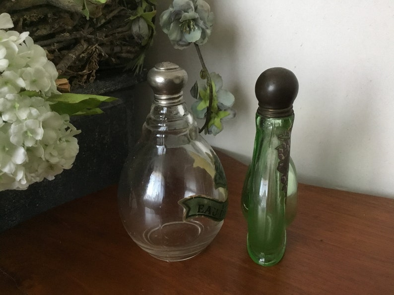 May include: Two vintage glass perfume bottles with silver tops. The bottle on the left is clear glass and has a label that reads "EAU DE". The bottle on the right is green glass.