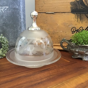 Könnte beinhalten: Eine gefrostete Glasbutterdose mit einem silbernen Deckel und einem dekorativen, geätzten Design. Die Dose steht auf einer Holzoberfläche.