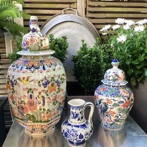 Könnte beinhalten: Eine Sammlung dekorativer Keramikvasen und ein kleiner Krug. Die Vasen sind weiß mit bunten Blumen- und Vogelmotiven und Deckeln. Der Krug ist blau und weiß mit floralen Mustern. Die Artikel werden auf einer reflektierenden Oberfläche ausgestellt.