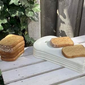 Op de afbeelding: Een stapel witte keramische broodborden met een enkele snee brood op elk bord. De borden zijn gestapeld op een witte houten tafel. Er is een stapel sneetjes brood aan de linkerkant van de afbeelding.