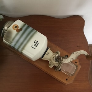 Könnte beinhalten: Vintage Wand-Kaffeemühle mit einem weißen Keramikbehälter mit der Aufschrift "Caffe" und einem Holzsockel. Die Mühle hat eine Metallkurbel und ein Glasgefäß zum Auffangen des gemahlenen Kaffees.