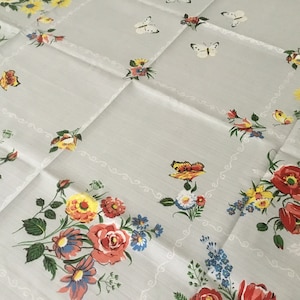 Könnte beinhalten: Eine weiße Tischdecke mit einem Blumenmuster mit roten, gelben und blauen Blumen und Schmetterlingen. Die Tischdecke hat eine zarte weiße Spitzenborte.
