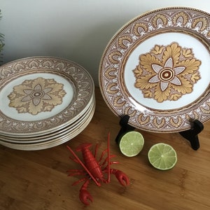 Könnte beinhalten: Ein Stapel von sechs weißen Keramiktellern mit einem braunen und goldenen Blumendesign. Die Teller sind auf einer Holzoberfläche gestapelt. Auf der Oberfläche befinden sich ein roter Hummer und zwei Limetten.