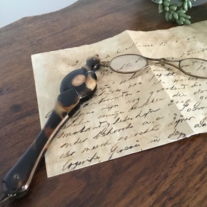 Könnte beinhalten: Ein Paar antike Klappbrillen aus Schildpatt mit einem handgeschriebenen Brief auf einer Holzoberfläche. Die Brille hat einen goldenen Metallrahmen und ist geschlossen. Der Brief ist in Kursivschrift geschrieben und datiert auf den "12. März 18..."