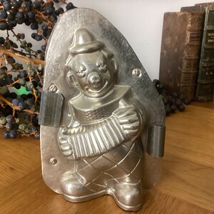 May include: A vintage metal mold for making chocolate shaped like a clown playing an accordion. The mold is silver in color and has a detailed design.