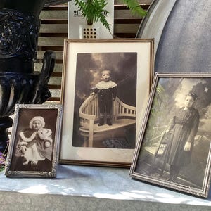 Könnte beinhalten: Drei antike gerahmte Fotografien. Das größte hat einen goldenen Rahmen und zeigt ein kleines Kind, das auf einer Bank steht. Die anderen beiden haben silberne Rahmen, eines zeigt ein Kind und das andere eine Frau. Die Fotos werden auf einer grauen Oberfläche ausgestellt.