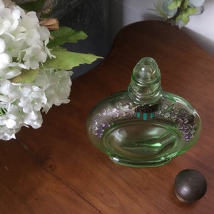 May include: A green glass bottle with a floral design and a clear glass stopper. The bottle is sitting on a wooden surface.