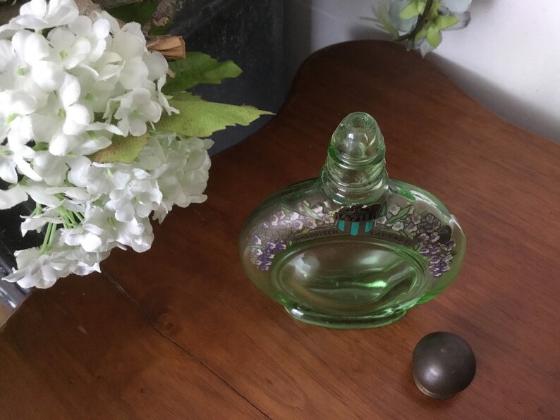 May include: A green glass bottle with a floral design and a clear glass stopper. The bottle is sitting on a wooden surface.