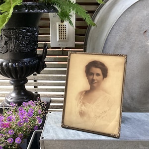 May include: An antique framed portrait of a woman in sepia tones. The portrait is in a gold-colored frame with ornate detailing. The image is set on a metal surface, with a black urn and purple flowers in the background.