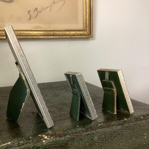 May include: Four silver picture frames with ornate designs, standing on a green surface. The frames are different sizes and are all angled to show the back.