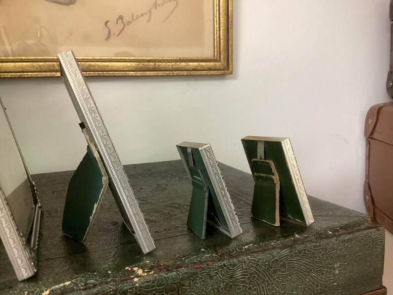 May include: Four silver picture frames with ornate designs, standing on a green surface. The frames are different sizes and are all angled to show the back.