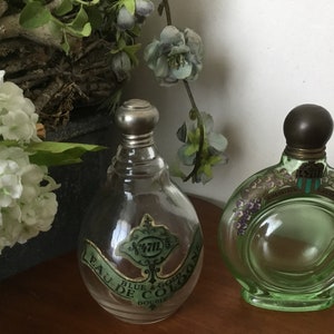 May include: Two vintage glass perfume bottles with silver and brass stoppers. The bottle on the left is clear glass with a green and gold label that reads "Eau de Cologne Blue & Gold Double", the bottle on the right is green glass with a white label that reads "Parfumerie Lavandou" and has a floral design.