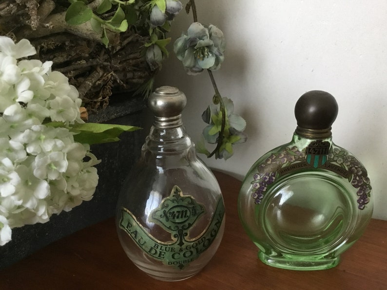 May include: Two vintage glass perfume bottles with silver and brass stoppers. The bottle on the left is clear glass with a green and gold label that reads "Eau de Cologne Blue & Gold Double", the bottle on the right is green glass with a white label that reads "Parfumerie Lavandou" and has a floral design.