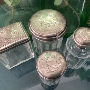 Könnte beinhalten: Vier durchsichtige Glasgefäße mit silbernen Deckeln. Jeder Deckel hat ein verziertes Design mit einem zentralen Oval.