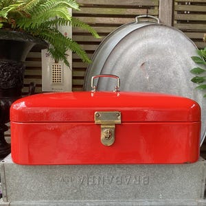 Può includere: Una cassetta degli attrezzi rossa brillante con chiusura e maniglia dorate. Poggia su un contenitore di metallo zincato. Sullo sfondo un vaso nero decorativo e vegetazione. La cassetta misura circa 30 cm di lunghezza.