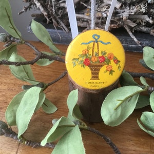 May include: A yellow round tin with a floral design and a blue ribbon. The tin has the text "HOUBIGANT PARIS" on the bottom.