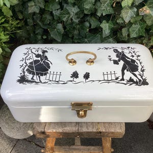 Puede incluir: Caja de pan de esmalte blanco con un diseño de silueta negra de dos figuras y un patrón floral. La caja tiene una manija de latón y un pestillo de latón.