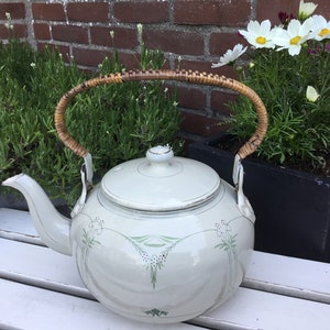 May include: A white enamel teapot with a green floral design and a woven wicker handle. The teapot is sitting on a white wooden surface.