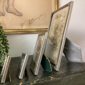 May include: Four silver picture frames of varying sizes, with the largest frame on the right and the smallest on the left. The frames are all standing upright on a dark surface. The largest frame has a photo of a person inside.