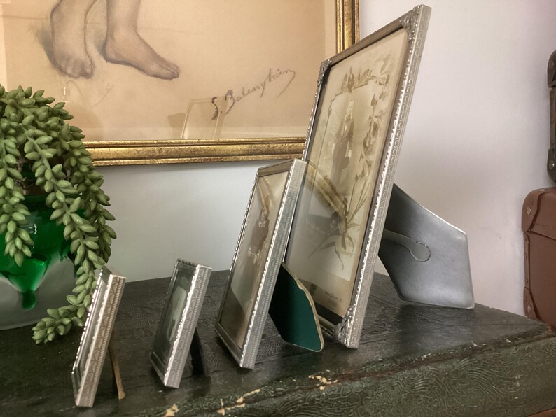 May include: Four silver picture frames of varying sizes, with the largest frame on the right and the smallest on the left. The frames are all standing upright on a dark surface. The largest frame has a photo of a person inside.