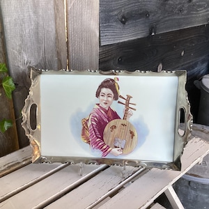 Può includere: Un vassoio rettangolare con una cornice in metallo color argento. Il vassoio presenta un'immagine dipinta di una donna in kimono che tiene uno strumento a corde. La donna è raffigurata in tonalità di rosso, marrone e beige.