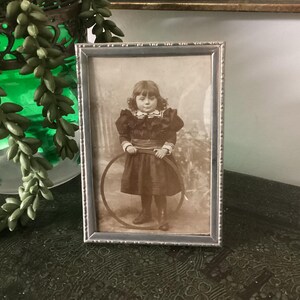 May include: A vintage silver-toned picture frame with a black and white photo of a young girl holding a hoop. The girl is wearing a dark dress with a white collar and a bow.