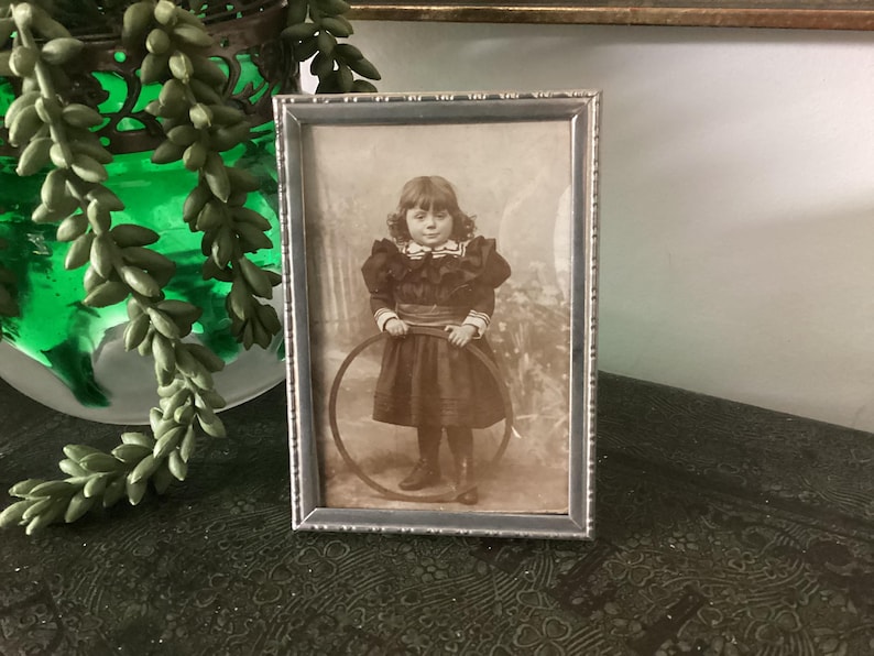 May include: A vintage silver-toned picture frame with a black and white photo of a young girl holding a hoop. The girl is wearing a dark dress with a white collar and a bow.
