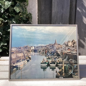 以下が含まれることがあります： 港の風景を描いたヴィンテージの額装されたプリント。運河に停泊しているボートが描かれている。プリントには、遠くの建物と教会の尖塔のある街が描かれている。空は白雲のある青い空である。