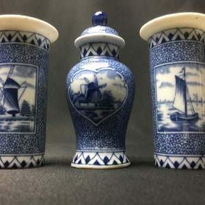 Op de afbeelding: Drie blauwe en witte keramische vazen met Nederlandse scènes. De middelste vaas is rond en bolvormig met een deksel. De twee zijvazen zijn cilindrisch. Alle drie vazen hebben een blauw en wit patroon met een windmolen en een boot.