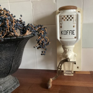 Peut inclure: Moulin à café en céramique blanche avec un dessus en bois et une poignée. Le moulin a un récipient en verre et le mot "Koffie" écrit sur le devant.