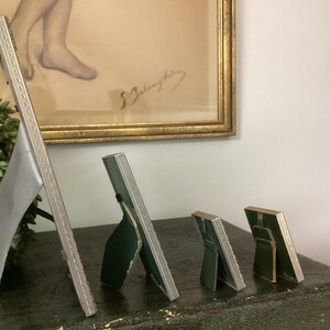 May include: Four silver picture frames of varying sizes, standing on a dark green surface. The frames are all ornate and have a silver finish. The largest frame is on the left and the smallest frame is on the right.