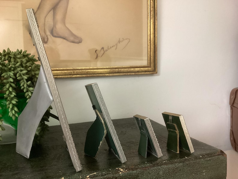 May include: Four silver picture frames of varying sizes, standing on a dark green surface. The frames are all ornate and have a silver finish. The largest frame is on the left and the smallest frame is on the right.