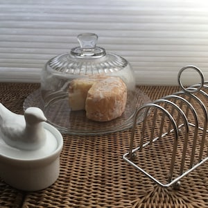 May include: A white ceramic bird-shaped butter dish with a lid, a glass cloche with a round cheese inside, and a silver metal bread knife holder on a woven wicker surface.