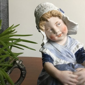 Könnte beinhalten: Eine Porzellanfigur eines jungen Mädchens, das ein blaues und weißes Kleid und eine weiße Haube trägt. Sie sitzt mit in den Schoß gelegten Händen und hat einen süßen Gesichtsausdruck.