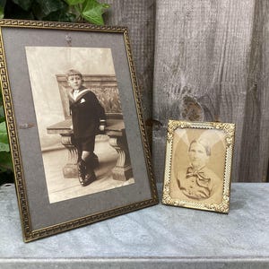 May include: Two antique picture frames. The larger frame holds a sepia-toned photograph of a young boy in a sailor suit. The smaller frame contains a portrait of a person with a bow tie. Both frames have ornate gold-colored borders.