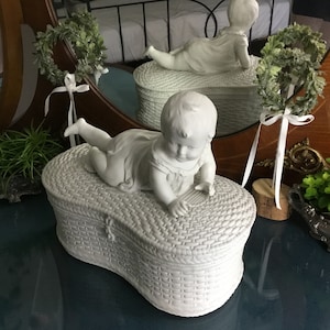 May include: White ceramic figurine of a baby lying on a white wicker basket with a lid. The baby is wearing a dress and has its arms outstretched. The basket is oval shaped and has a woven pattern.
