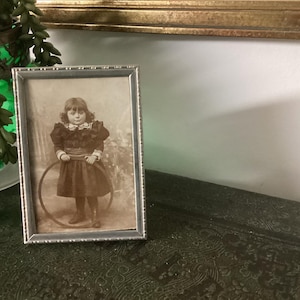 May include: A black and white photograph of a young girl holding a hoop. The photo is framed in a silver frame with a decorative border.