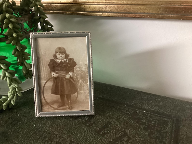 May include: A black and white photograph of a young girl holding a hoop. The photo is framed in a silver frame with a decorative border.