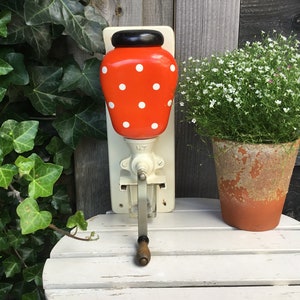 Peut inclure: Un moulin à café vintage rouge et blanc à pois avec un dessus noir et une poignée en bois. Le moulin est monté sur un mur blanc et possède une manivelle en métal.