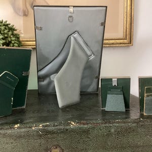 May include: Four silver picture frames with green felt backing. The frames are arranged in a row on a green surface. The largest frame is standing upright, while the three smaller frames are lying flat.
